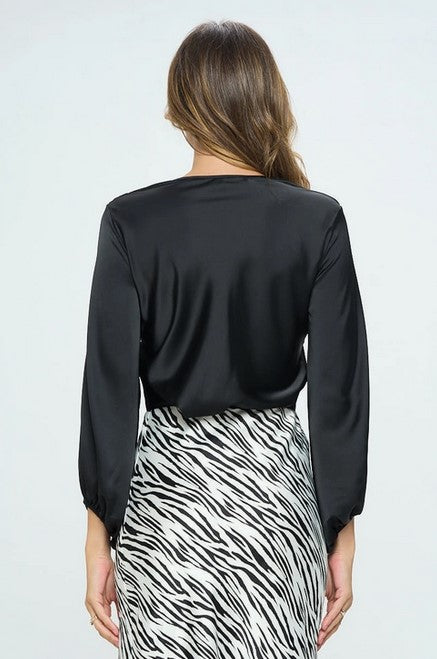 Black V-neck satin blouse with a twisted detail in the front. The model is wearing it tucked into a black and white zebra pattern skirt and is posing facing away from the camera in front of a white background.